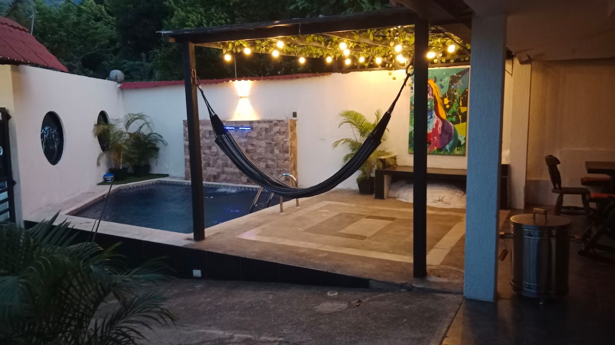 An outdoor area featuring a private pool, surrounded by palm plants and illuminated by string lights. A hammock is suspended nearby, and a colorful piece of wall art adds a vibrant touch. A wooden dining table and chairs are positioned on the patio.