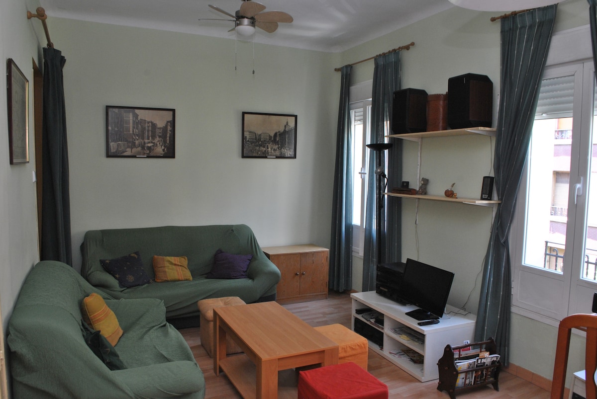 The living area features a light green color scheme, with two sofas in soft fabric and a wooden coffee table at the center. A TV is positioned on a low stand, accompanied by a multi-level shelving unit holding decorative items. Large windows allow natural light to enter the space.