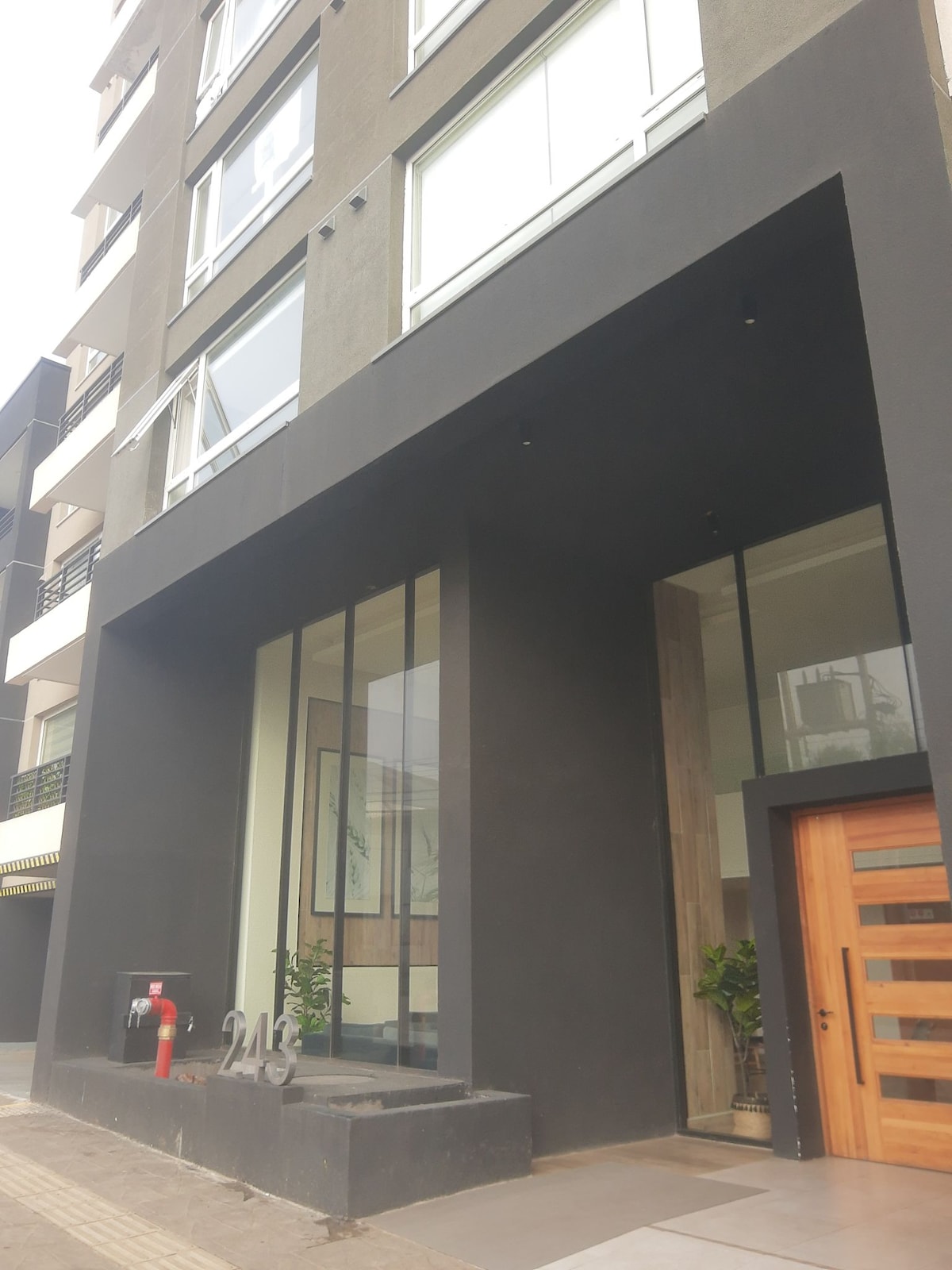 The entrance of a modern building features a large glass facade, providing a view inside. A combination of dark and light materials is used for the exterior, highlighted by a wooden door. The building number is prominently displayed at the base.
