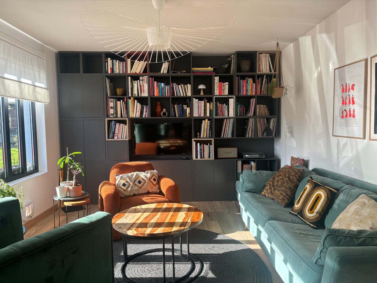A cozy living area is displayed, featuring a teal sofa and an orange armchair. A wooden coffee table rests in the center, surrounded by a bookcase filled with books. Natural light illuminates the space, highlighting the textured rug below.