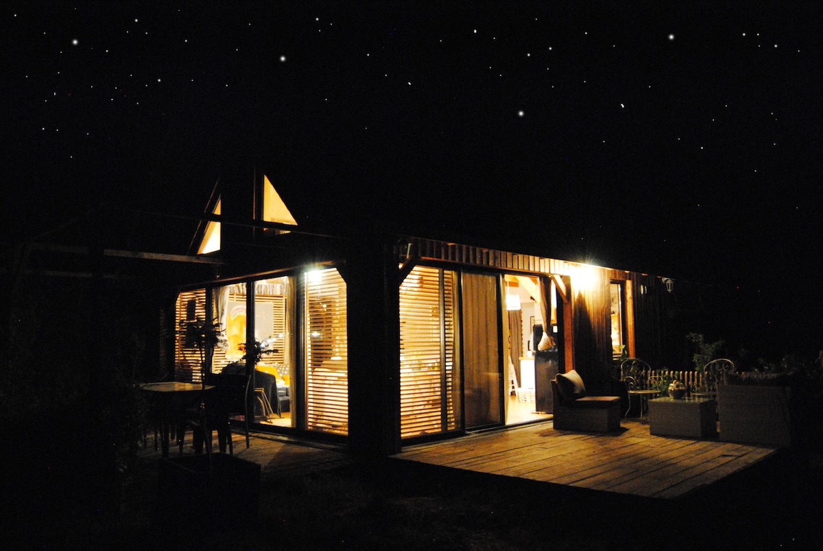 The house is illuminated at night, showcasing large glass doors that reveal the warm interior. Soft lighting highlights the wooden structure, while the starry sky adds a serene backdrop. Outdoor seating is visible on the deck, inviting relaxation in the evening.