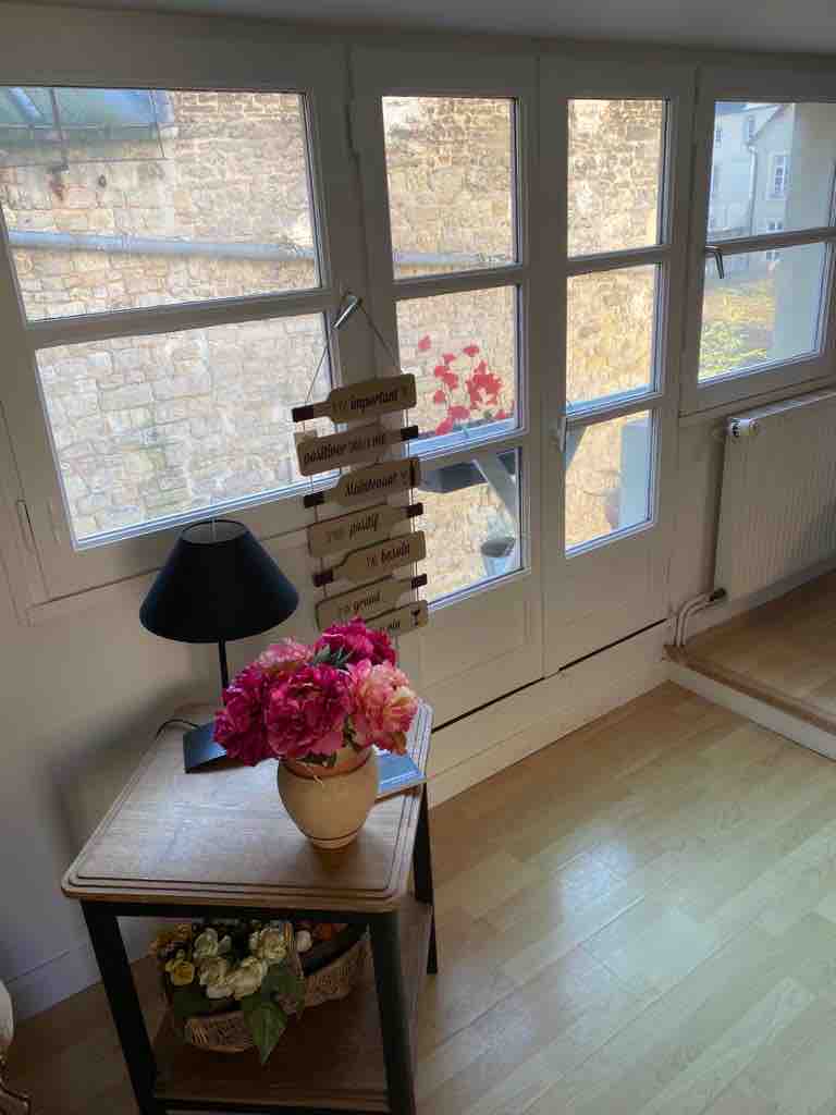 Large windows allow natural light to fill the space, revealing a view of historical stone walls. A lamp provides soft lighting beside a small table, which displays a decorative vase of colorful flowers and a quaint arrangement of dried plants.