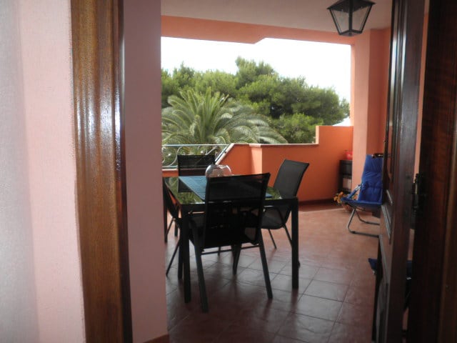 A covered terrace is visible, featuring a dining table with black chairs. Lush greenery is seen beyond the railing, contributing to a serene outdoor ambiance. The tiled floor adds a functional element to the space, while a small folding chair is positioned in the corner.