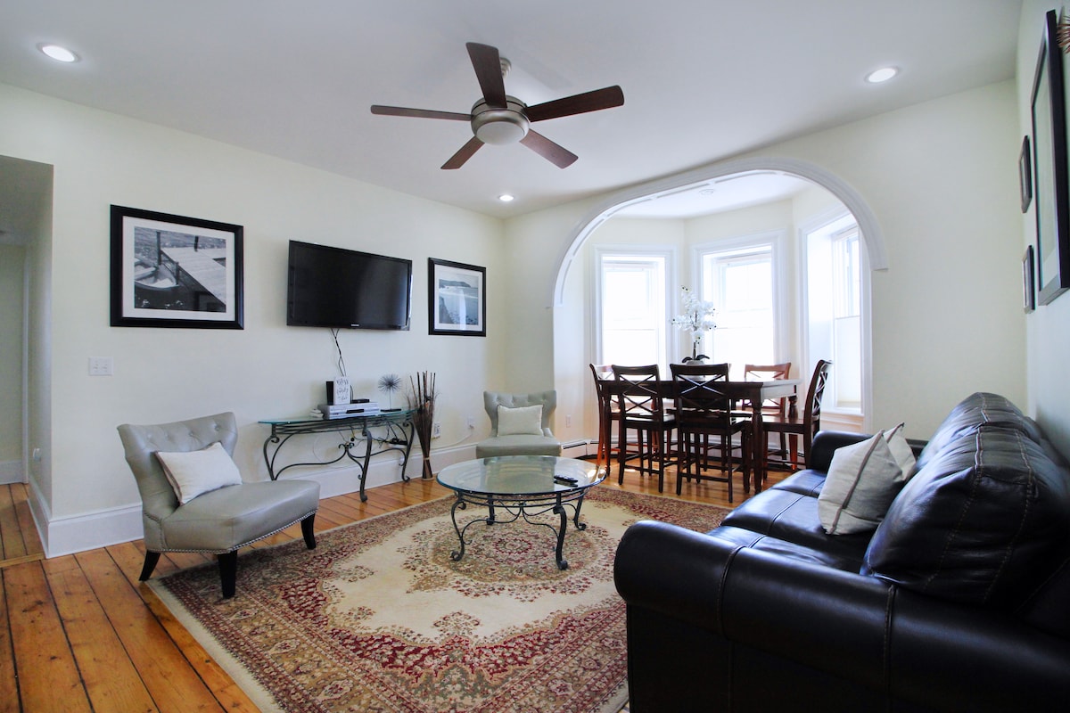 A well-lit living area features a comfortable leather sofa and two upholstered chairs arranged around a glass coffee table. A decorative rug adds warmth to the wooden floor, while a ceiling fan provides airflow. Nearby, a dining area with wooden chairs and a round table is flanked by large windows.