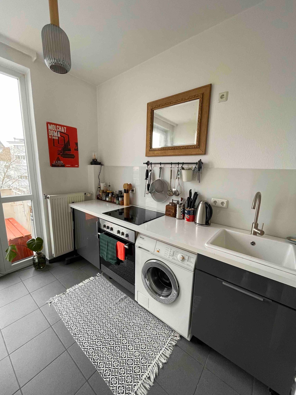 A modern kitchen setting is depicted, featuring sleek gray cabinetry and essential appliances, including a stove and washing machine. A small mirror is positioned above the sink, while a textured mat lies on the floor. A potted plant adds a touch of greenery near the window.