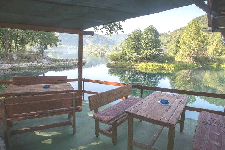 A wooden terrace overlooks the calm waters of the Una River, surrounded by lush greenery. Several rustic wooden tables and benches are arranged on the deck, creating a cozy outdoor seating area. The serene landscape is complemented by reflections of trees on the water's surface.