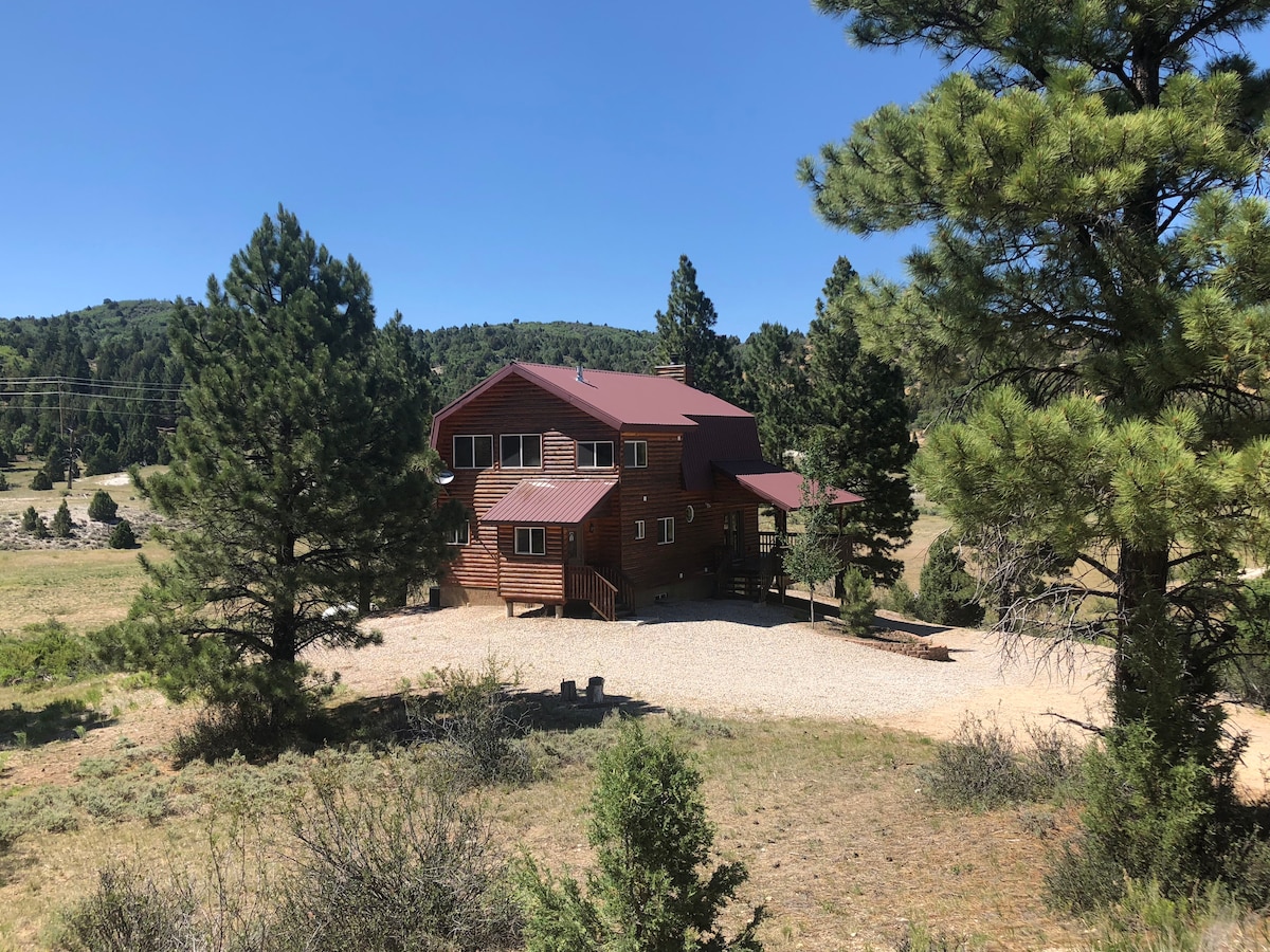 A spacious home with a rustic exterior is surrounded by a natural landscape of trees and grass. A gravel driveway leads to the entrance, while a covered deck extends around the building. Mountains are visible in the background under a clear blue sky.