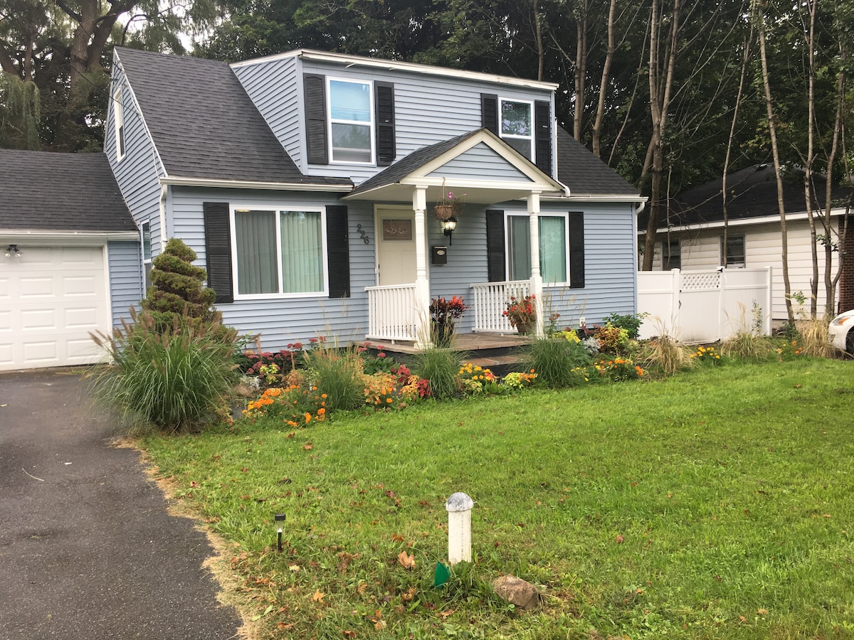 A charming two-story house features a light blue exterior and a welcoming front porch. A well-maintained garden with various colorful flowers surrounds the entrance, enhancing the home's curb appeal. The driveway leads to a garage visible on the left side.