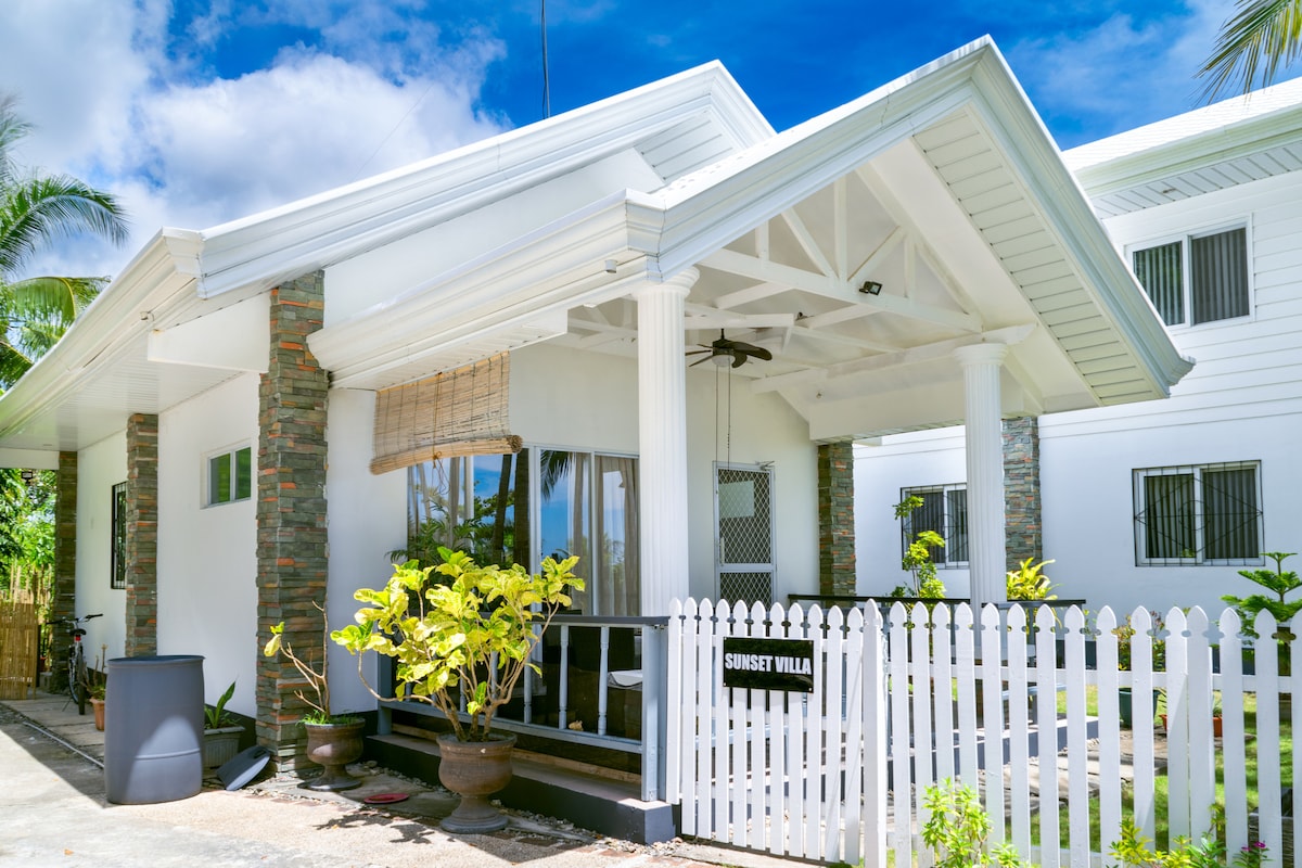 The exterior of Sunset Villa is showcased, exhibiting a modern design with a white facade and stone accents. The entrance features a covered porch adorned with tropical plants and a welcoming sign. A white picket fence encloses the area, enhancing the privacy and charm.
