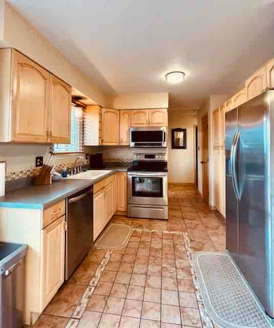 A fully stocked kitchen is shown, featuring light wood cabinetry and modern stainless steel appliances. Counter space is ample, with a sink positioned below a window. Tile flooring is present, complemented by decorative mats in front of the appliances.