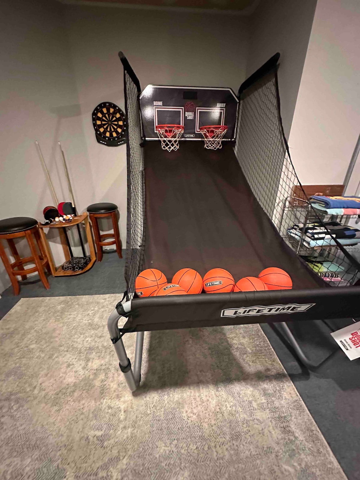 An indoor basketball shooting game is showcased, featuring two hoops positioned at the top of a sloped ramp. Several orange basketballs are neatly arranged at the bottom of the ramp. Bar stools and a dartboard are visible in the background, enhancing the entertainment space.
