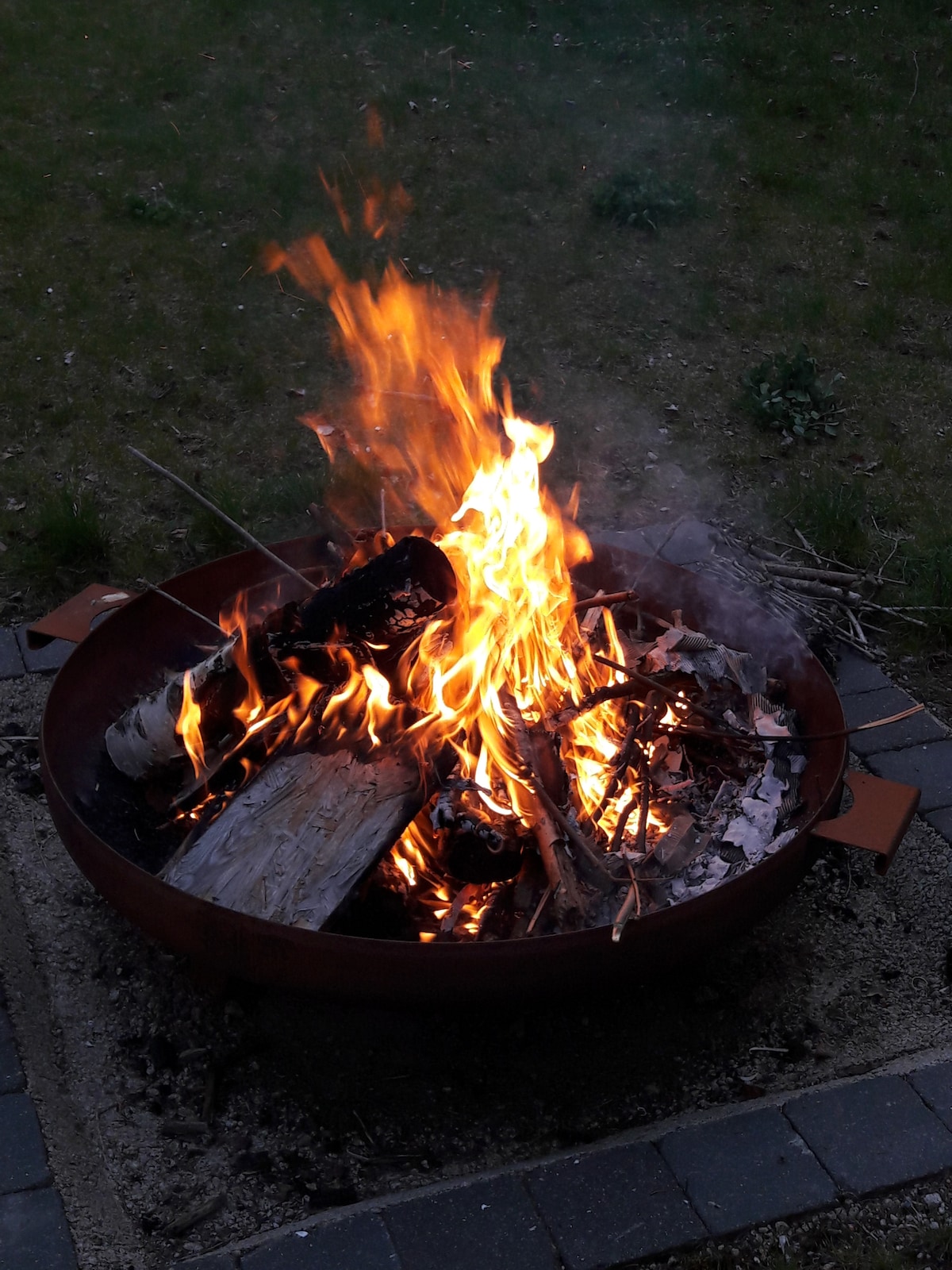 A fire burns brightly in a circular metal fire pit, surrounded by small stones. Flames dance above a collection of logs and twigs, creating a warm glow against the evening backdrop. The grassy area surrounding the fire pit offers a natural setting for relaxation.