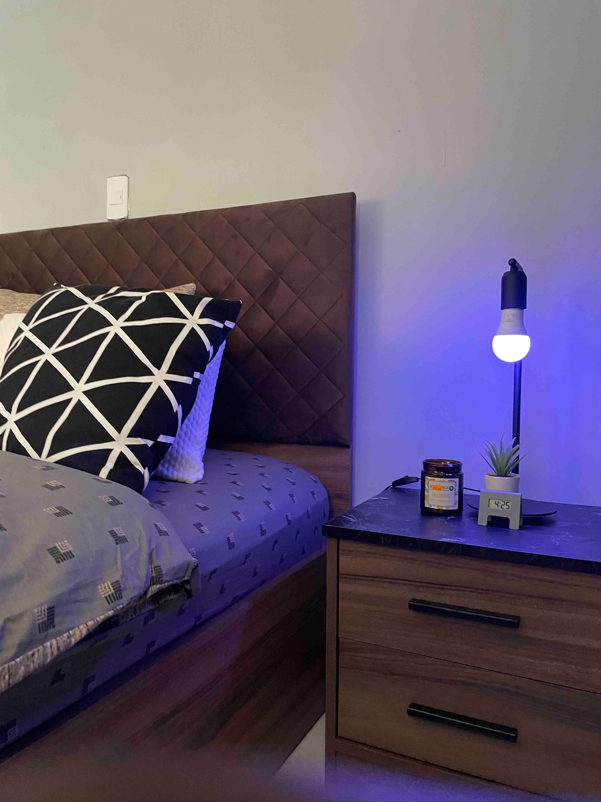 A spacious bed features an elegant upholstered headboard and is adorned with a patterned blanket. A bedside table holds a small lamp with a blue light and a potted plant, alongside a jar. Subtle textures and colors create a calming atmosphere.