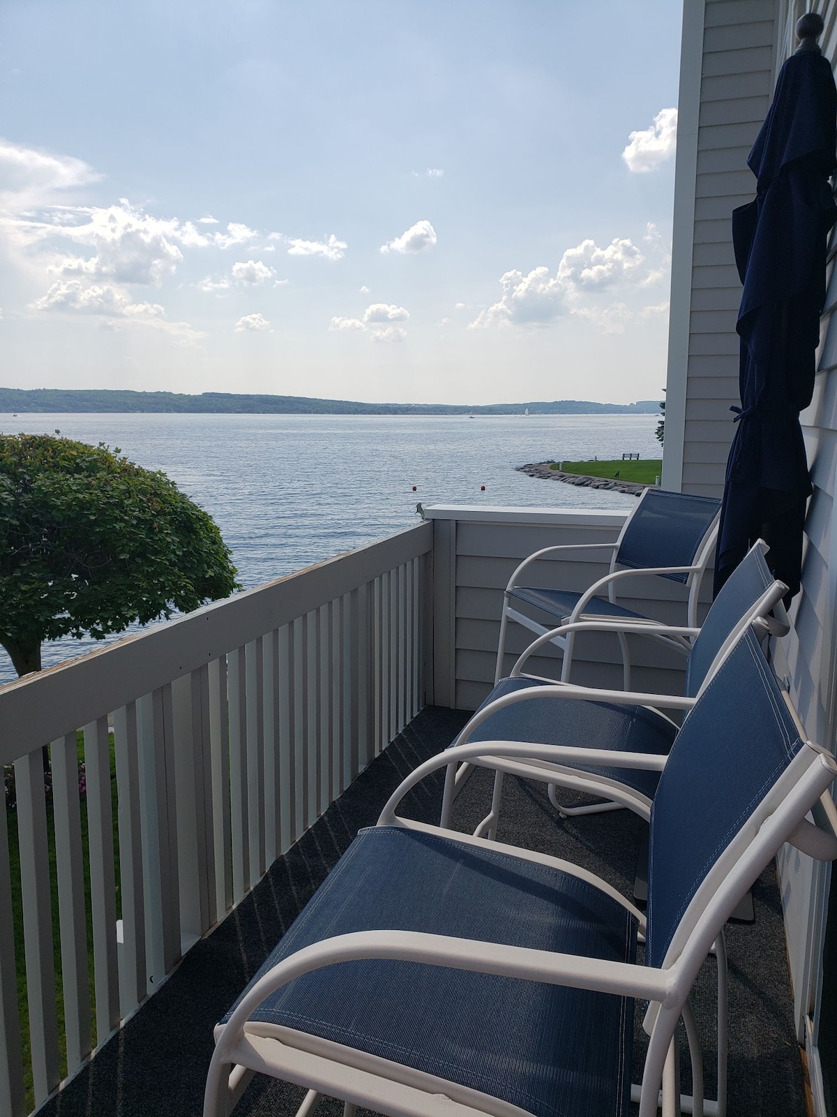 A balcony is equipped with four white chairs featuring blue cushions, offering views of the calm waters of Lake Charlevoix. The scene is framed by a railing and a nearby tree, with the distant shoreline visible under a partly cloudy sky.