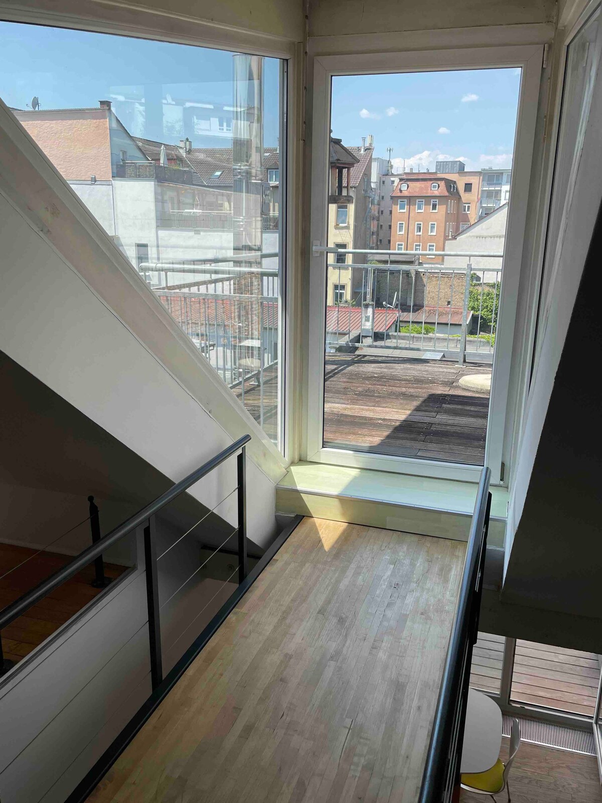 A view from a second-floor gallery showcases a bright and airy space. Large glass windows allow natural light to flood the area, providing a glimpse of the surrounding cityscape and a private terrace below. The sleek railing adds a modern touch to the overall design.