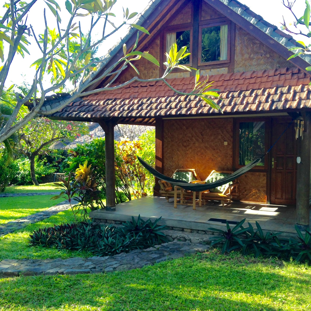 A traditional bungalow is positioned within a lush tropical garden. The entrance features a porch with two comfortable chairs and a hammock, surrounded by vibrant greenery. Wide windows allow natural light to fill the space, offering a serene outdoor view.