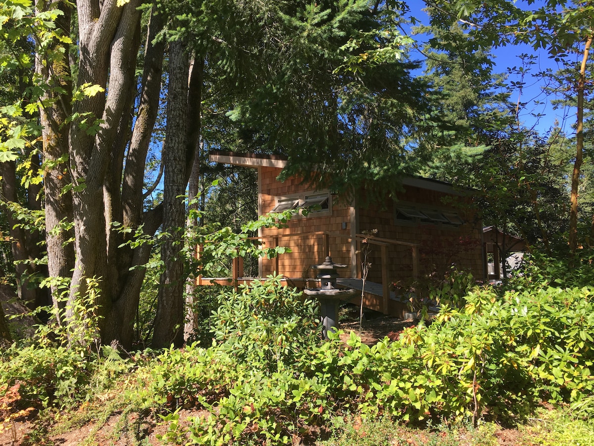 A cozy cottage is nestled among trees, with cedar shingles and a metal roof visible. Surrounding greenery includes lush shrubs and tall trees, creating a tranquil forest ambiance. Soft sunlight filters through the foliage, illuminating the rustic exterior and inviting porch space.