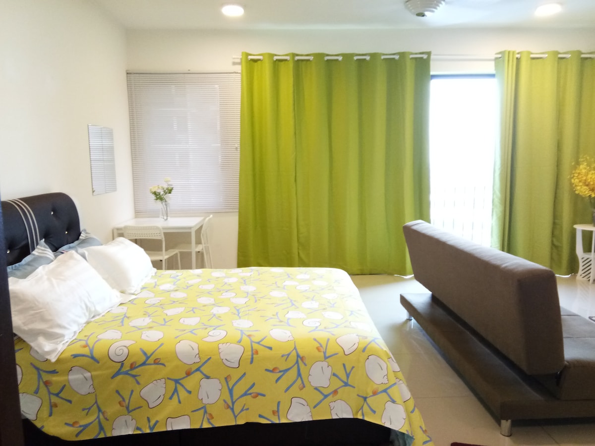 A spacious bedroom features a large bed adorned with a bright yellow bedspread patterned with sea shells. A comfortable sofa is positioned nearby, and large green curtains frame the window, allowing natural light to fill the room. A small side table holds a vase with fresh flowers.