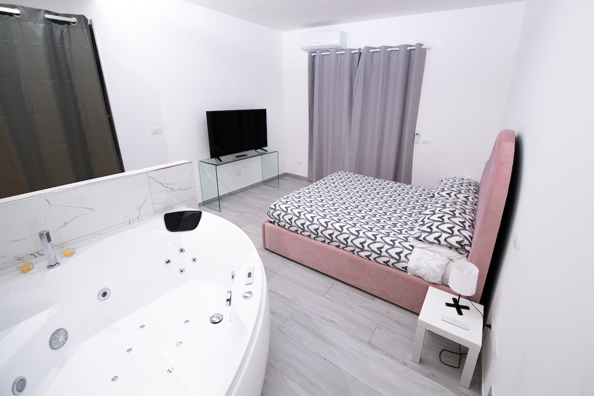 A spacious room is presented, featuring a luxurious jacuzzi and a cozy bed with patterned bedding. A flat-screen TV is mounted on a glass stand, and curtains partially cover a window, allowing for some natural light to enter the space.