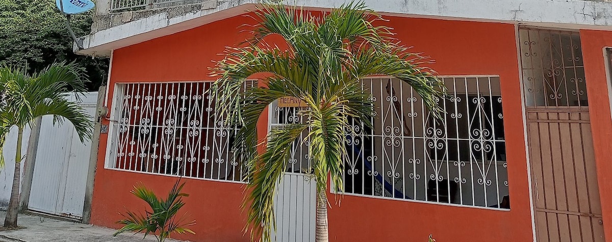A vibrant orange exterior features a welcoming entrance with a white door. The front is framed by grassy landscaping and palm trees, complemented by decorative wrought iron grilles on the windows. Natural light illuminates the area, enhancing the inviting atmosphere of the property.
