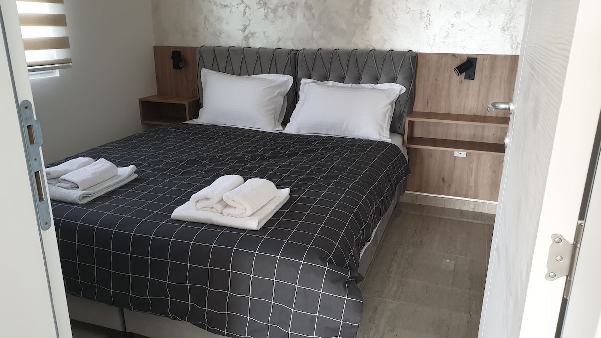 A spacious sleeping area features a neatly made bed with two white pillows and a black and white checkered blanket. Soft white towels are placed at the foot of the bed. Simple wood nightstands with wall-mounted reading lights complement the modern interior design.