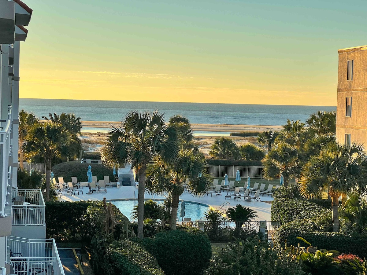 A view from the third-floor balcony showcases the beach and ocean in the distance, framed by palm trees. The serene pool area is visible in the foreground, surrounded by lounge chairs and lush greenery, all bathed in the warm glow of a sunset.