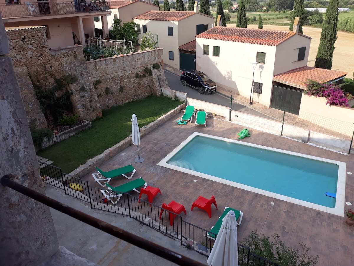An aerial view of a shared outdoor area is displayed, featuring a rectangular swimming pool surrounded by lounge chairs. Red and green reclining chairs are positioned around the pool, while two white umbrellas provide shade. Lush greenery and a stone wall complete the serene setting.