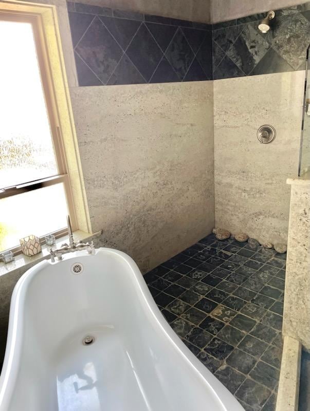 A bathroom is shown featuring a clawfoot bathtub and a walk-in shower. Natural light enters through a window, illuminating the stone tile design. The shower is adorned with smooth river rocks, providing a unique aesthetic. The walls display a mix of textured stone and tiles.