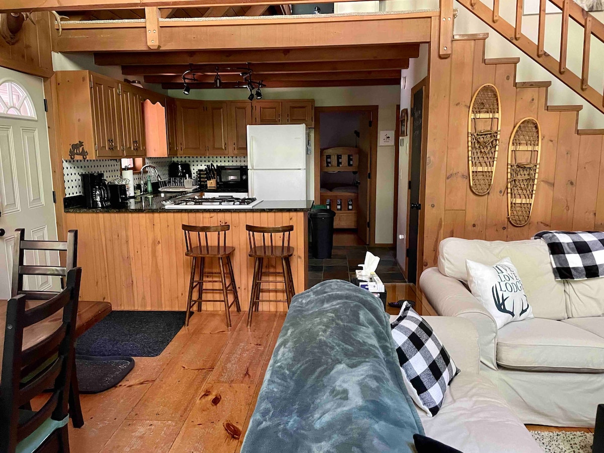 An open-concept kitchen and living area features wooden cabinets and a spacious island with two stools. A comfortable seating area includes a light-colored couch and a coffee table. Natural wood elements and a cozy atmosphere are highlighted throughout the space.