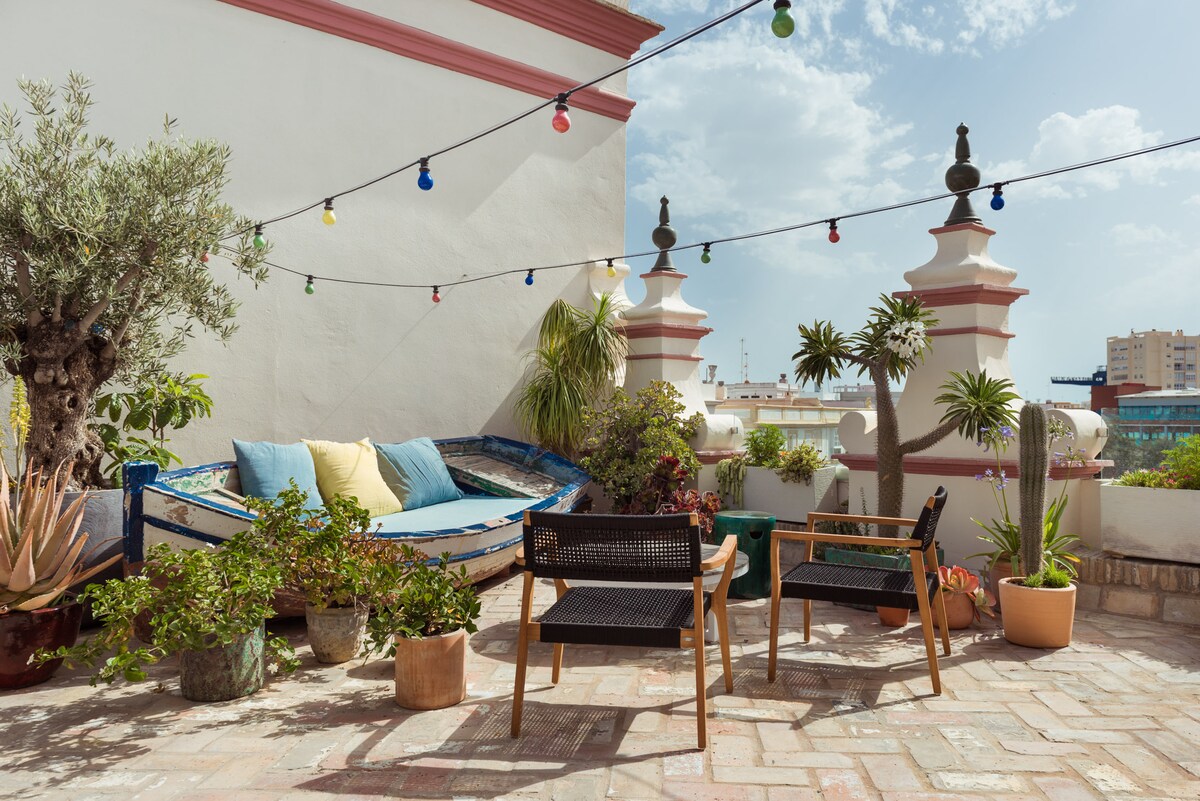 A rooftop terrace features a variety of potted plants and colorful lights strung above. Two black chairs and a cozy blue seating area invite relaxation. Sunlight illuminates this tranquil space, surrounded by decorative walls and distant city views.