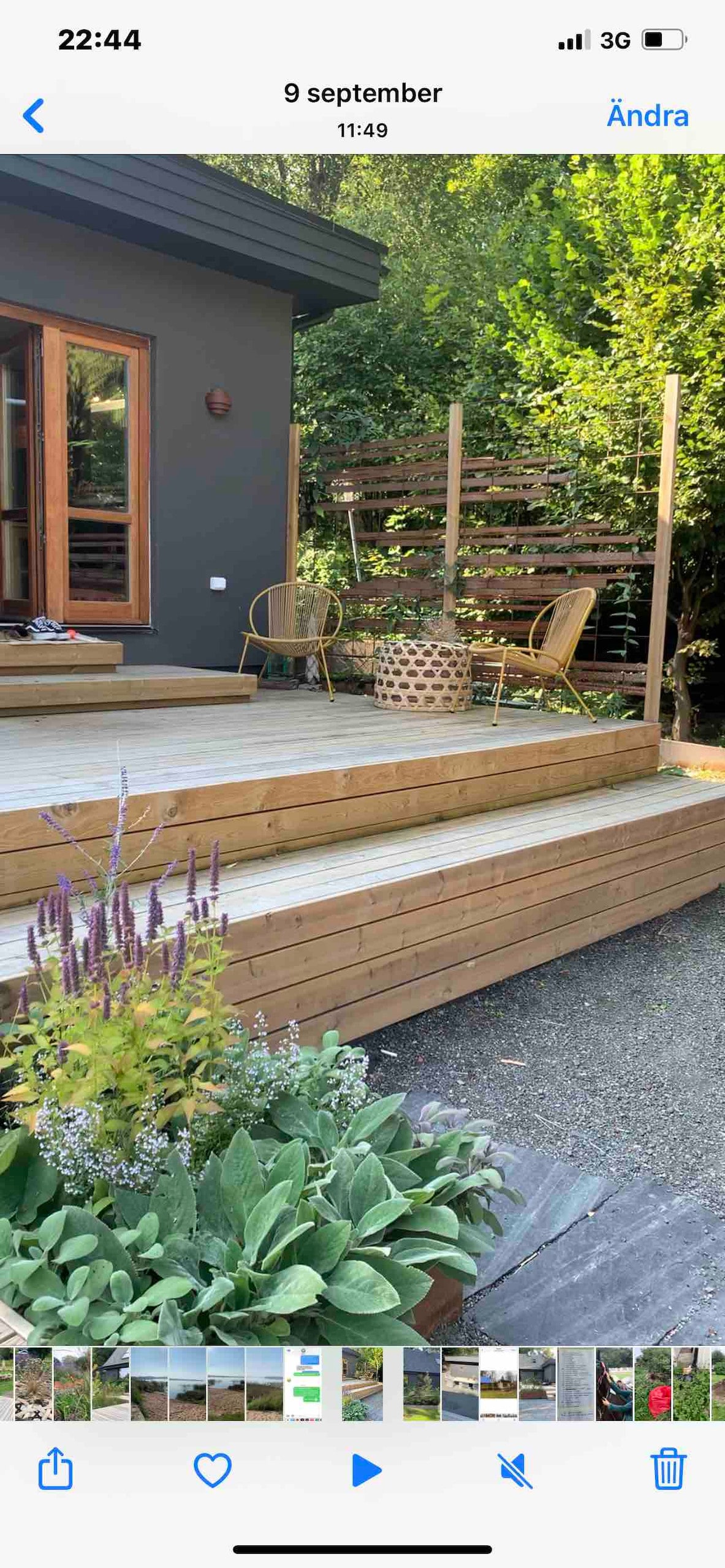 An outdoor wooden deck features steps leading to a cozy seating area with rattan chairs. Lush greenery surrounds the space, complemented by potted plants with colorful flowers. Natural light highlights the warm tones of the deck, inviting relaxation in a serene environment.