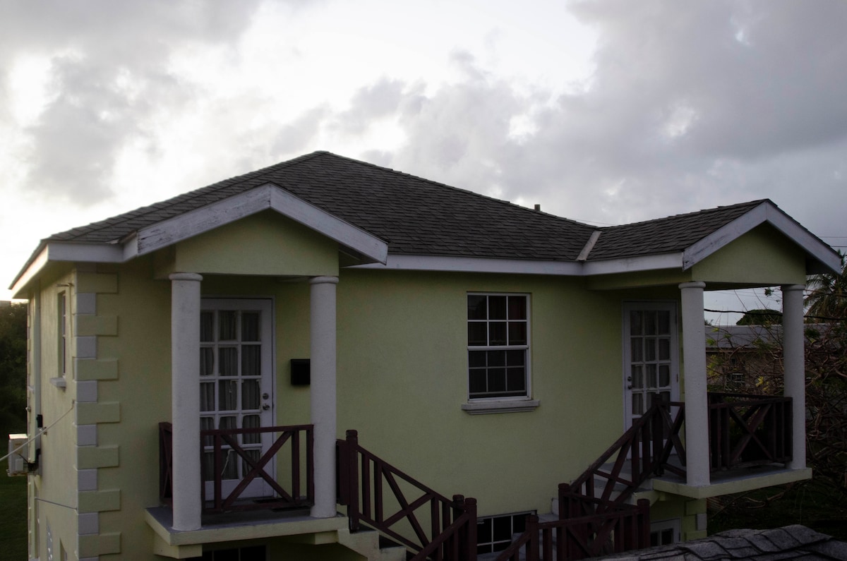 The exterior of the two-story apartment features a light green facade with a gabled roof. Sturdy wooden railings adorn the front steps and balconies, while large windows provide ample natural light. A cloudy sky adds a serene backdrop to the structure.