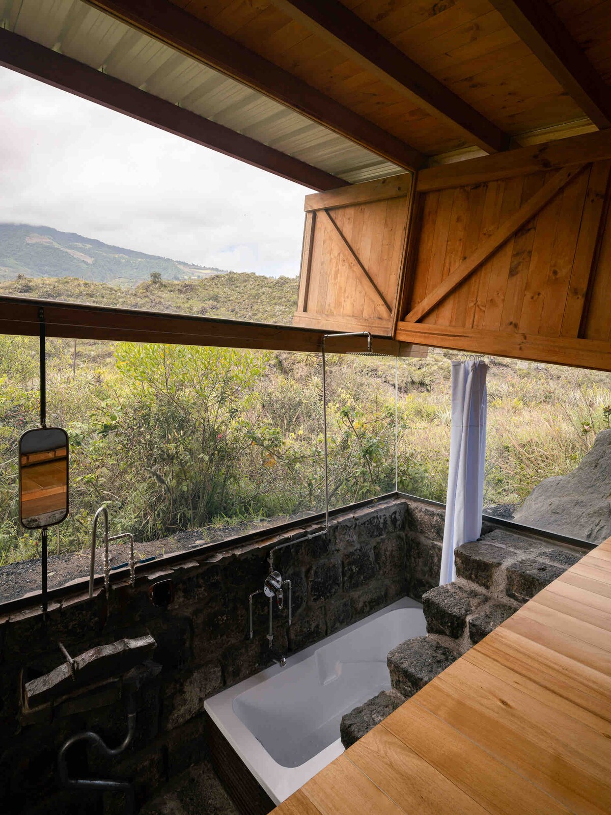 A private bathroom features a modern bathtub positioned near glass walls, offering scenic views of the surrounding landscape. Natural light floods through the angled wooden ceiling, illuminating the stone and wood accents that create a serene atmosphere.