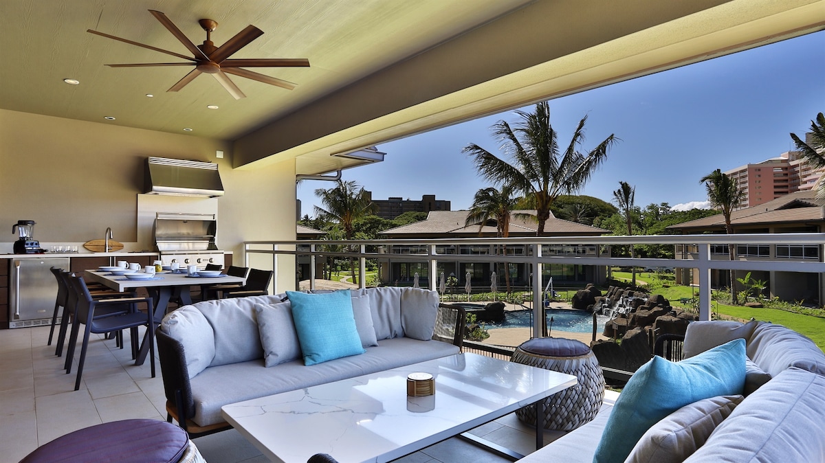 The spacious lanai hosts a comfortable seating area with a coffee table and decorative accents. A dining table is positioned nearby, complemented by an outdoor kitchen. Palm trees and manicured grounds are visible beyond, providing a backdrop of tropical greenery and the pool area.