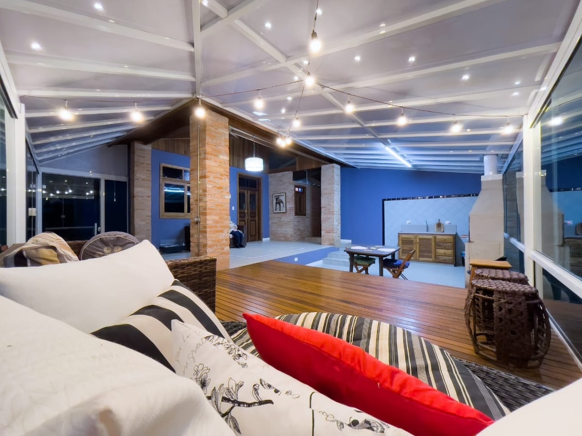 An outdoor deck area is illuminated by string lights, featuring cozy seating with decorative pillows. A view of a spacious living room with a fireplace and stylish decor is visible through large glass doors. The ambiance is welcoming, perfect for relaxation or gatherings.
