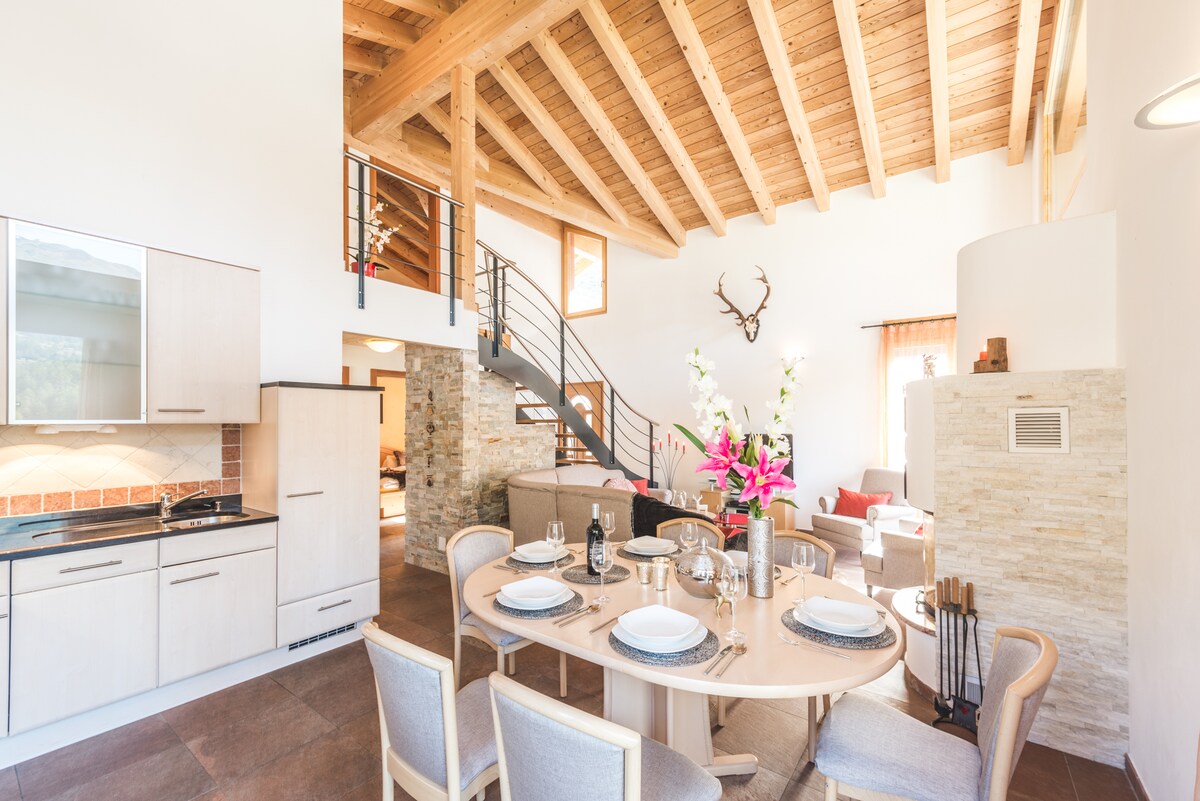 An open-plan kitchen and dining area is showcased with a round wooden table set for a meal. A modern kitchen with light cabinetry is seen, along with attractive stone accents on the walls. A spiral staircase leads to an upper level, and wooden beams are visible on the ceiling.