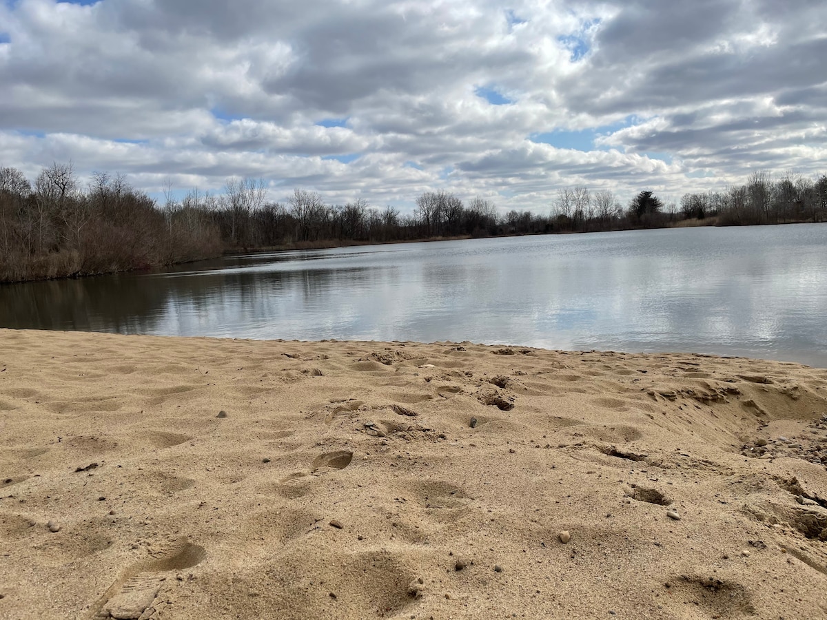 A sandy beach area extends towards a tranquil lake, reflecting cloudy skies. The shoreline features footprints in the sand, with sparse vegetation lining the water's edge. The calm lake is surrounded by trees, enhancing the natural setting.