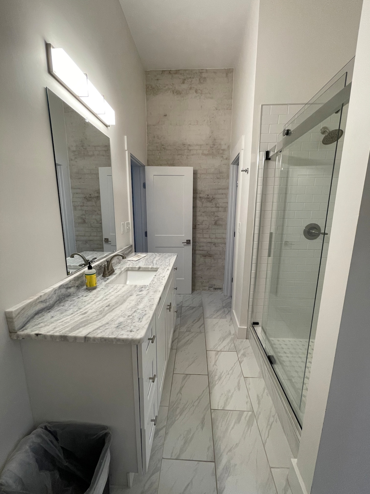 A modern bathroom is presented, featuring a sleek double vanity with a marble countertop. A large mirror is mounted above the sinks, and a glass-enclosed shower is visible at the end of the room. The floor is tiled with light-colored marble, enhancing a spacious feel.