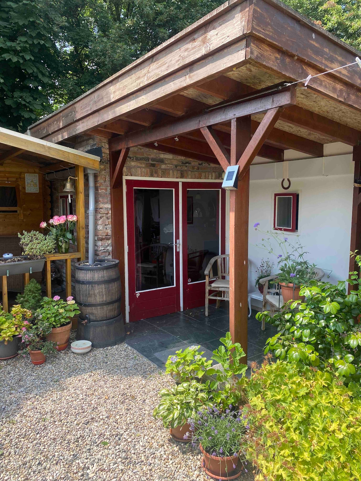 An entrance area is framed by a wooden overhang. The red double doors provide access to the interior, while potted plants and flowers are arranged along the perimeter. A rustic barrel and additional greenery enhance the welcoming ambiance of the space.