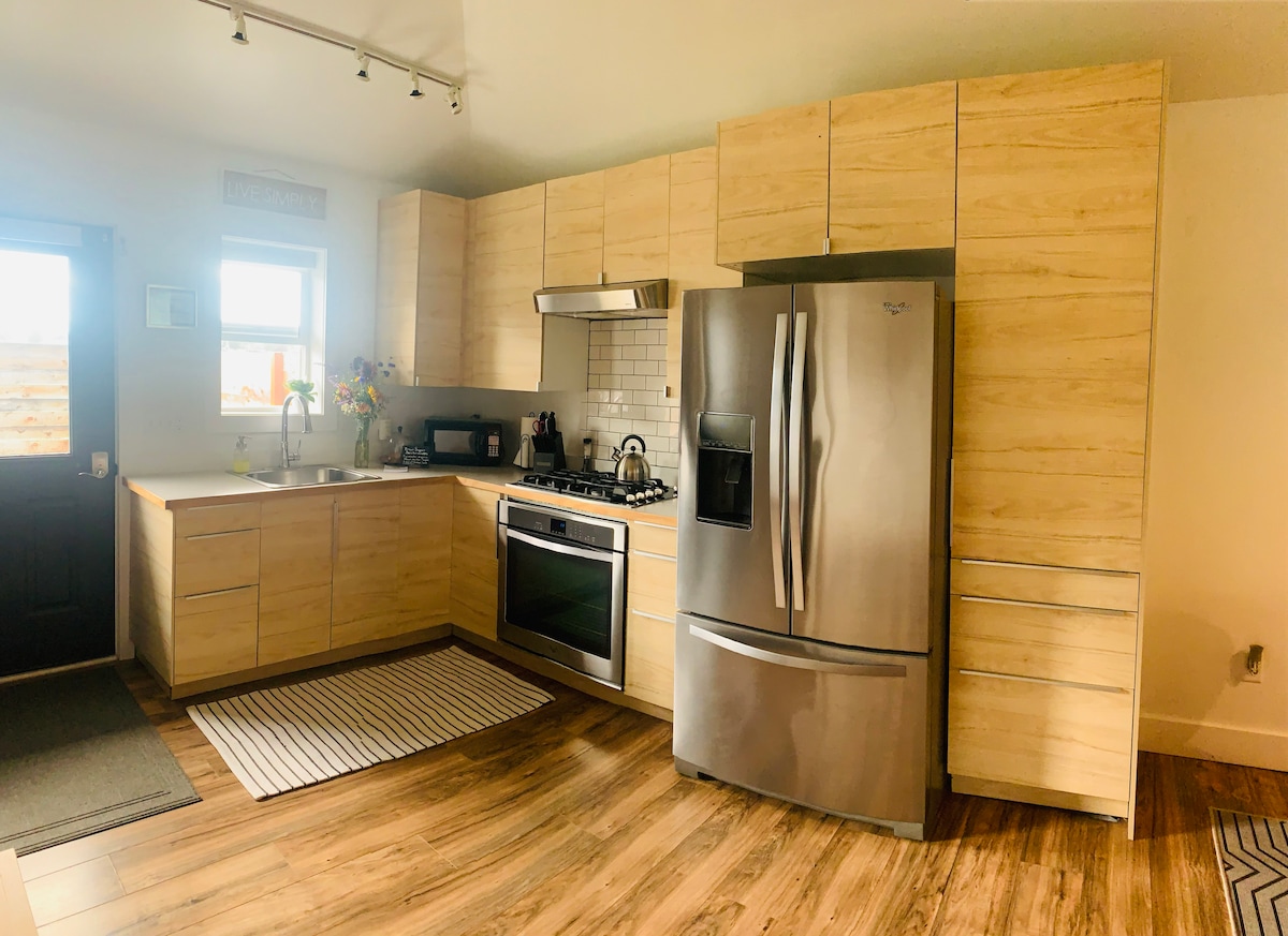 A modern kitchen is featured, showcasing light wood cabinetry and stainless-steel appliances. The space includes a gas stove and a large refrigerator. A subtle area rug lies on the floor, and a door leads outside, allowing natural light to brighten the room.