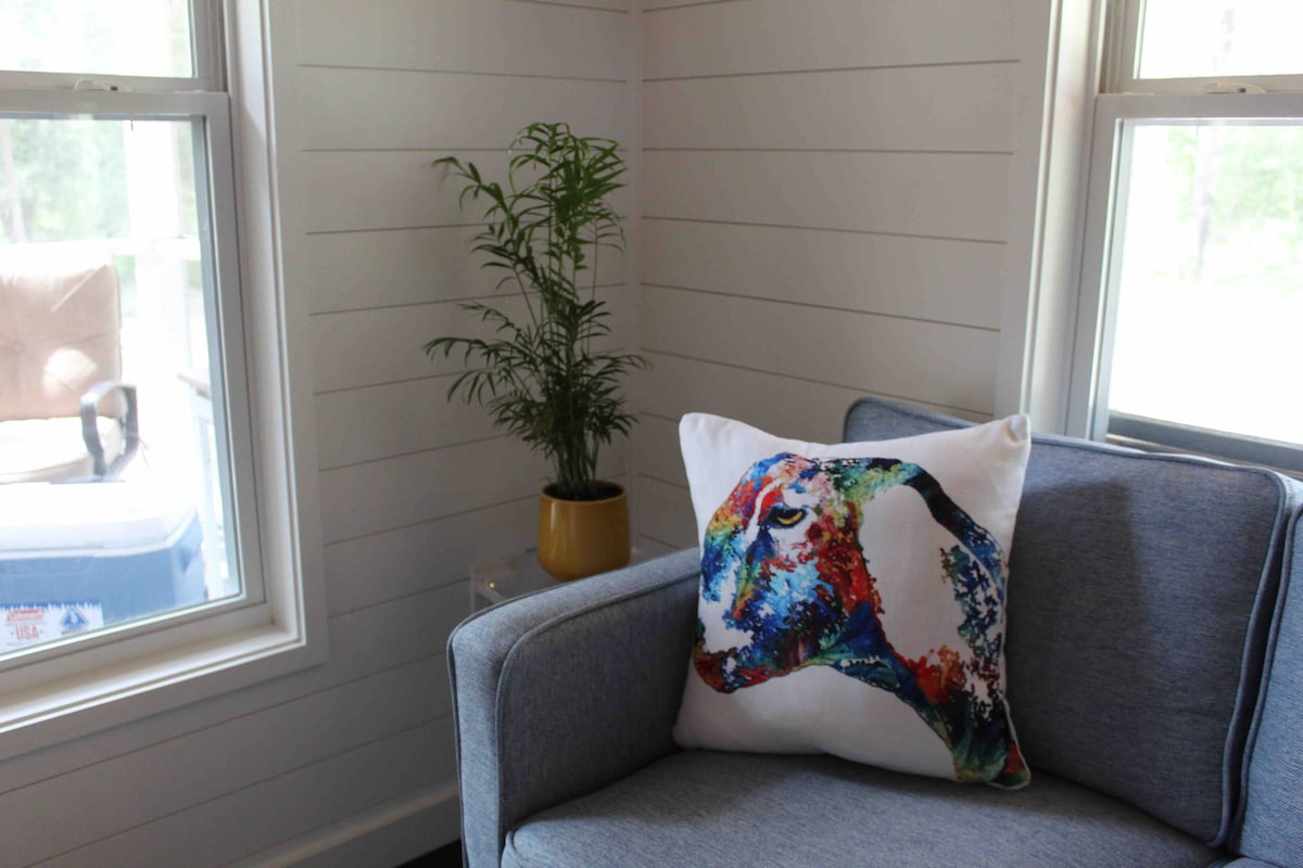 A cozy corner features a light gray sofa adorned with a vibrant throw pillow displaying a colorful animal design. A lively green plant is positioned in a yellow pot on a nearby windowsill, with natural light streaming in through two windows.