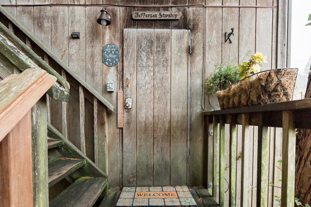 The entrance features a wooden door with a rustic finish, adorned with a welcome mat and a sign labeled 'Jefferson Starship.' Potted plants are positioned alongside the door, enhancing the natural aesthetics of the wooden staircase leading up to the entryway.