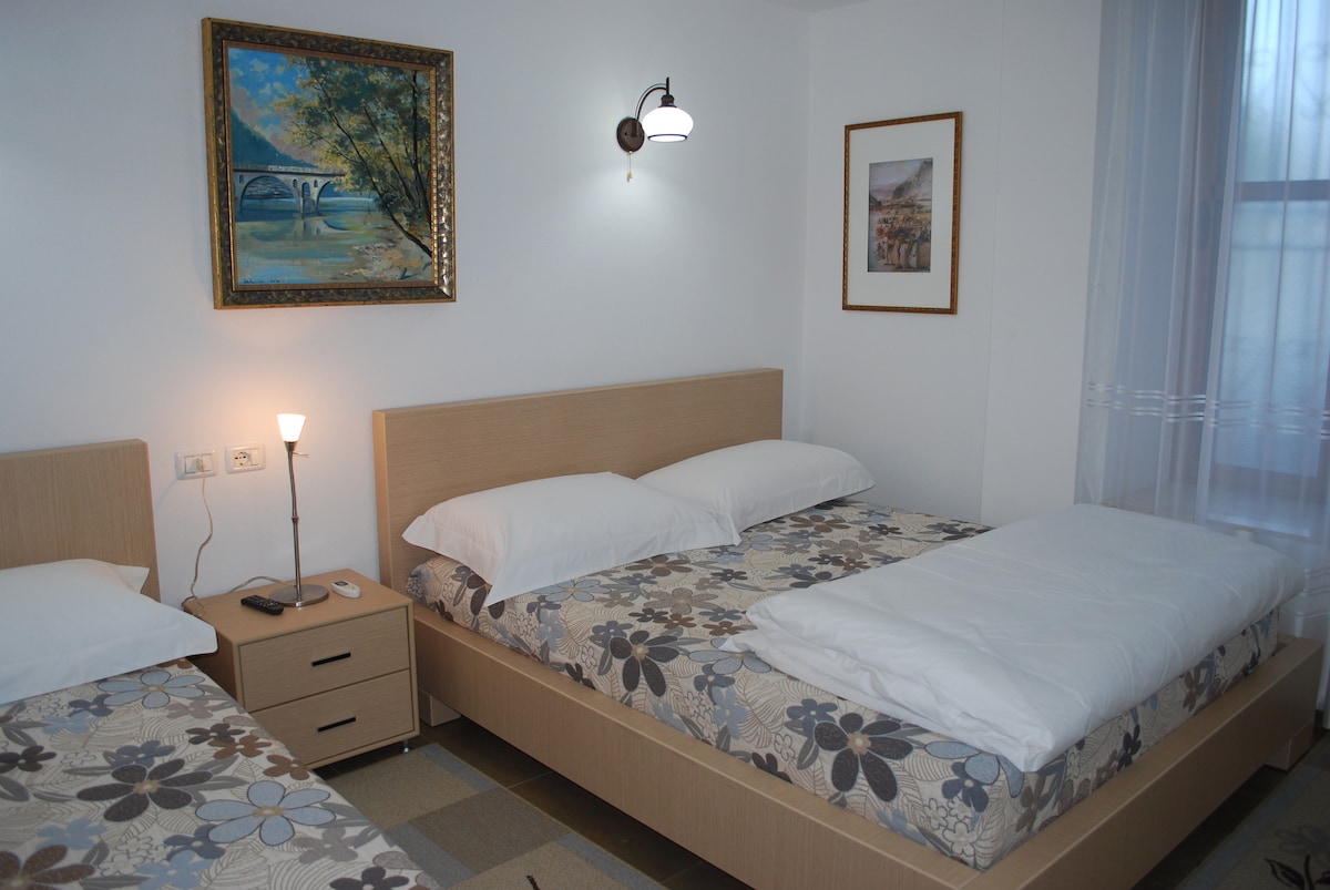 A well-lit, airy room features two spacious beds with floral-patterned bedding. A bedside lamp casts a warm glow, and artwork adorns the walls. Natural light streams in through a window with sheer curtains, creating a bright and inviting atmosphere.