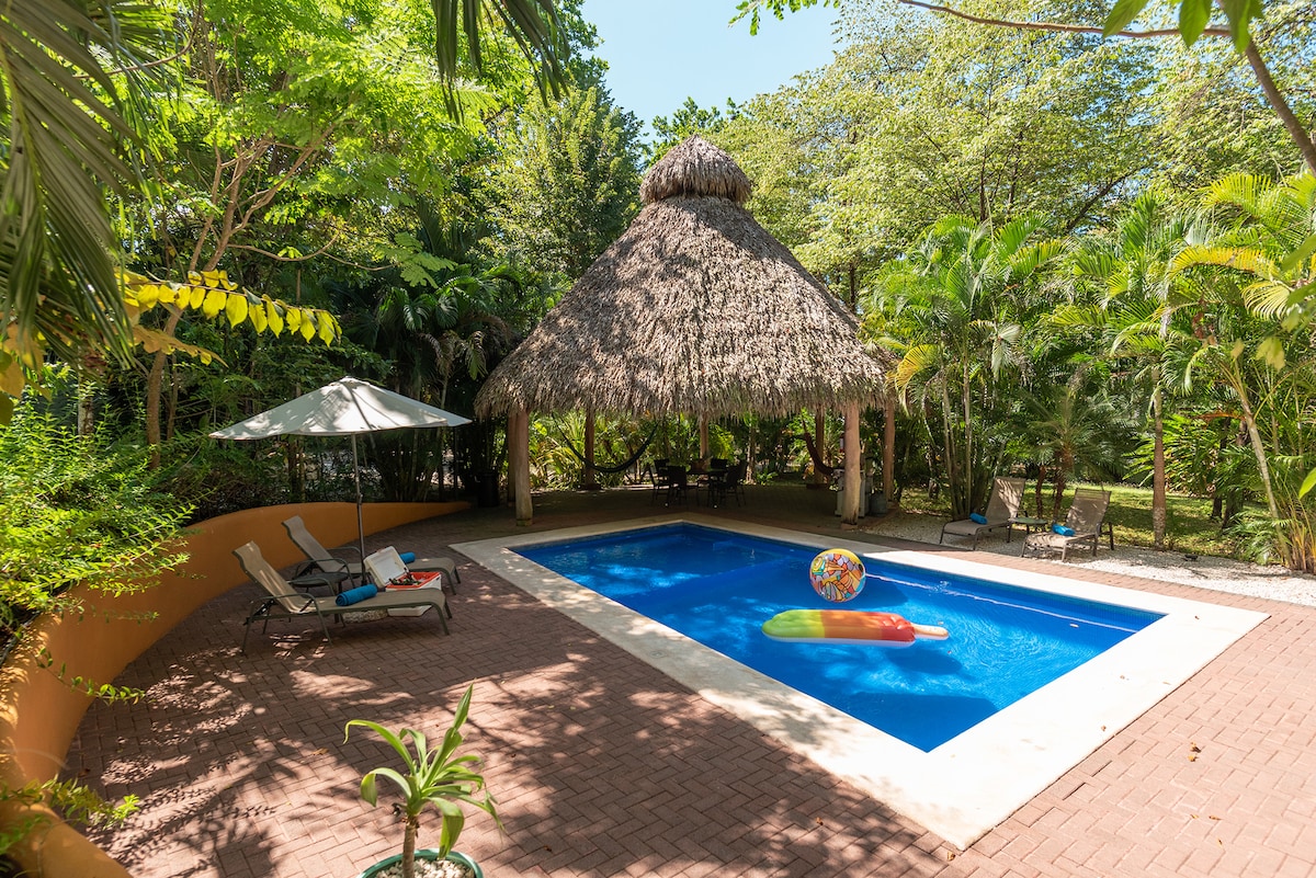 A shaded outdoor area features a pool surrounded by a spacious deck of laid bricks. A straw-roofed tiki hut provides cover, while sun loungers sit nearby. A colorful beach ball floats in the inviting blue water, complemented by lush greenery that frames the scene.