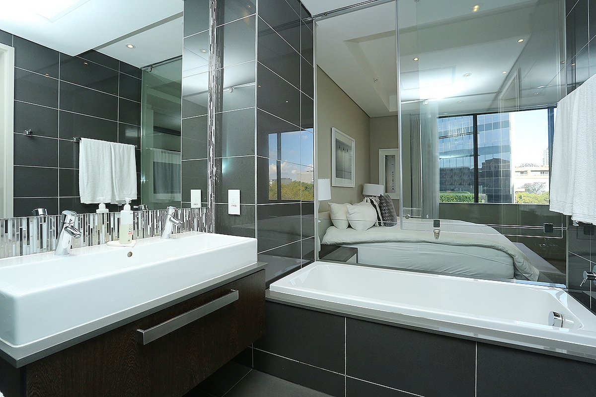 A modern bathroom features a sleek double sink vanity with a spacious countertop and ample storage. A large mirror reflects the light from the window, creating an open feel. A white bathtub is positioned next to the shower area, enhancing the contemporary design.