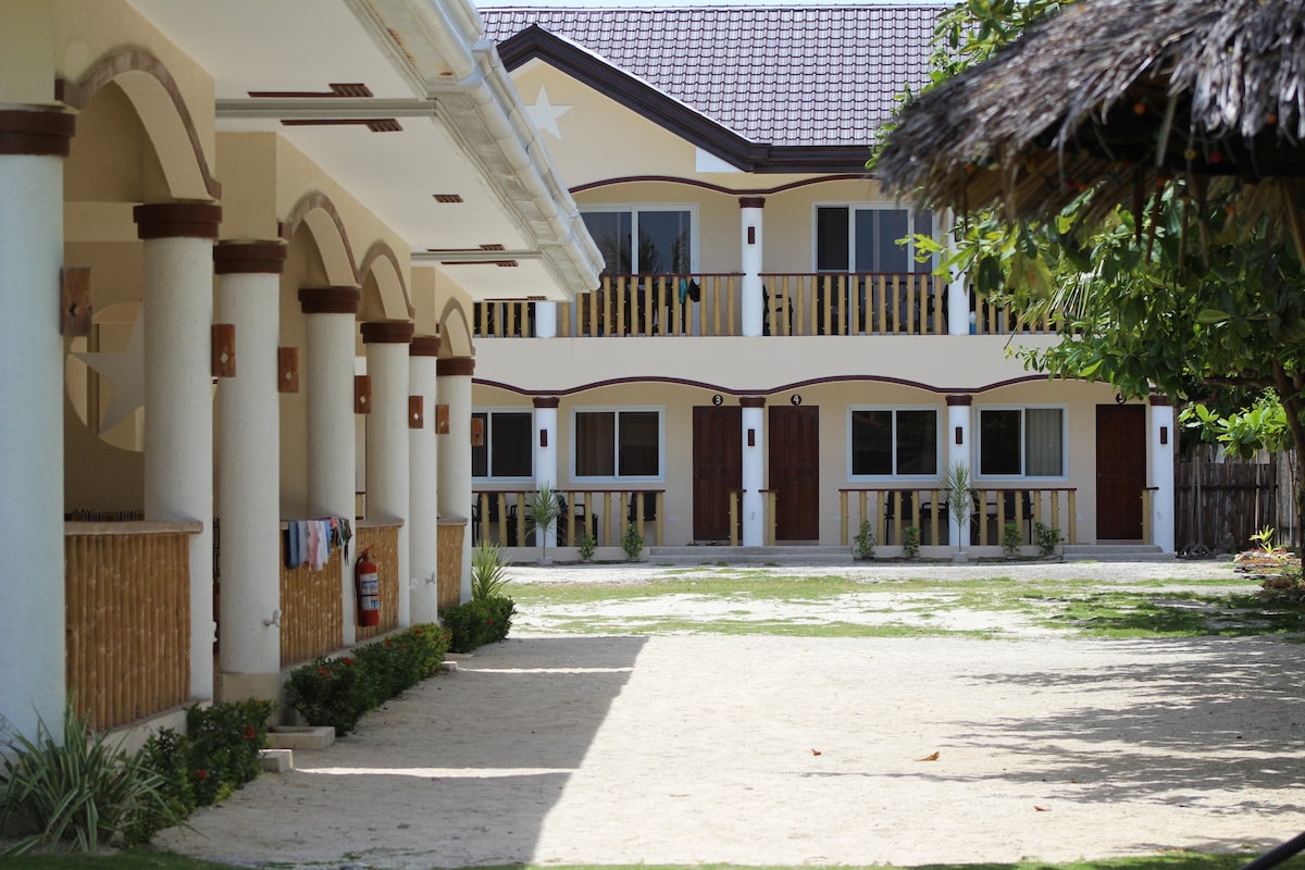 A spacious courtyard is shown, featuring several arched columns leading to a building with wooden doors and large windows. Lush greenery surrounds the sandy area, creating a serene environment. The arrangement of the buildings offers ample space for outdoor relaxation.