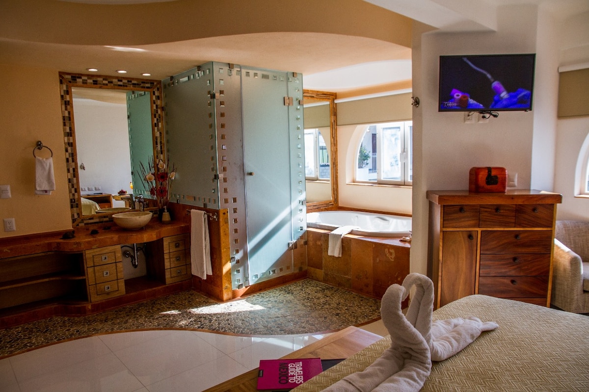 A spacious bathroom features a glass-enclosed shower and a jetted tub. Natural light streams through multiple windows, illuminating the warm color palette. Wooden cabinetry and unique tiling enhance the elegant design. A flat-screen television is mounted on the wall, providing entertainment options.