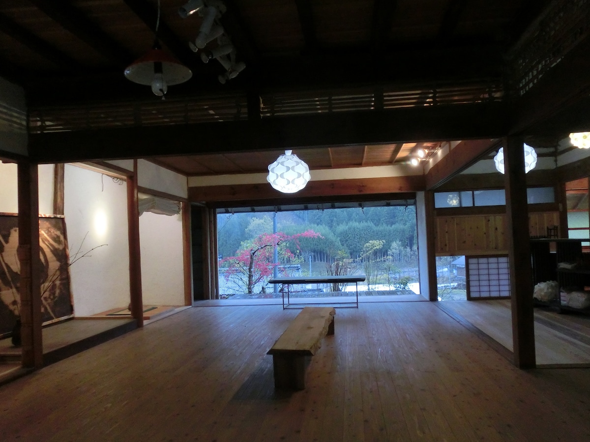 A spacious interior features polished cedar floorboards and is illuminated by soft pendant lighting. Large glass doors open to reveal a serene view of the outdoors, where vibrant trees can be seen. A simple wooden bench sits centrally in the room.