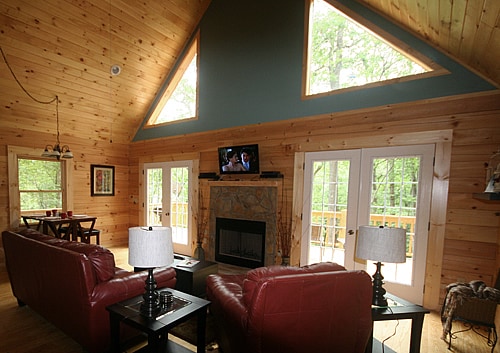 The living area features vaulted ceilings and large triangular windows, allowing natural light to illuminate the wooden interior. Two leather sofas are positioned around a stone fireplace, with a television mounted above. Two side tables with lamps are placed beside the sofas, creating a cozy atmosphere.
