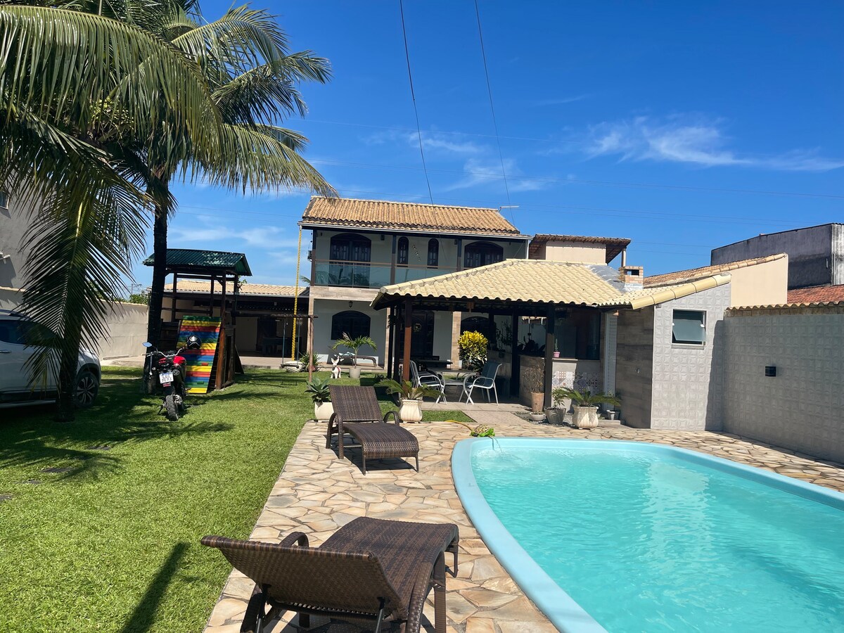 A spacious outdoor area is visible, featuring a turquoise pool surrounded by lounge chairs on a stone patio. A shaded gazebo offers seating options, while palm trees provide greenery. The house's upper level can be seen in the background, reflecting a warm, inviting design.
